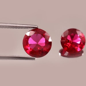 May include: Two round, faceted, bright red gemstones. One is held by silver tweezers, the other rests on a reflective surface. The gems have a brilliant cut, showcasing their internal facets and vibrant color. The background is a soft, neutral tone.