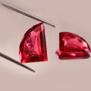 May include: Two faceted, semi-circular, bright red gemstones. One is held by tweezers, the other rests on a reflective surface. The gems have multiple facets that catch the light, creating a sparkling effect. The background is a soft, neutral color.