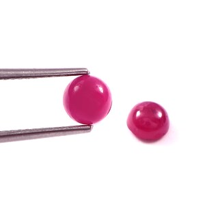 Peut inclure: Deux petites pierres précieuses rondes taillées en cabochon d'une couleur rose magenta vive et unie. Une pierre est maintenue fermement entre les pointes métalliques d'une pince argentée, tandis que la seconde pierre identique est posée à côté.
