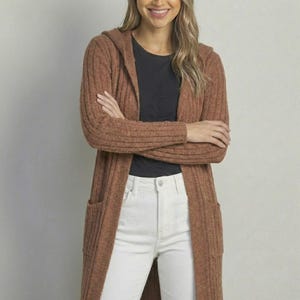 May include: A long, hooded cardigan in a warm brown color. The sweater has a ribbed texture, two front pockets, and a relaxed fit. The model is wearing a black top and white jeans.