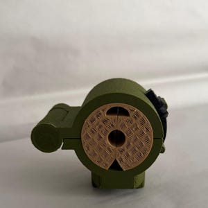 May include: A 3D-printed, olive green object with a cylindrical body and a circular, textured face. The face is a light brown color. A small black component is attached to the side. The object has a mechanical appearance.