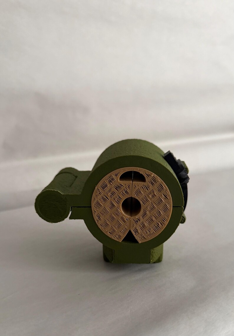 May include: A 3D-printed, olive green object with a cylindrical body and a circular, textured face. The face is a light brown color. A small black component is attached to the side. The object has a mechanical appearance.