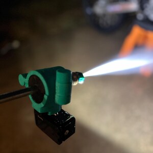 May include: A close-up of a green and black pressure washer nozzle emitting a bright, white stream of water. The nozzle is mounted on a metal rod. The background is dark and out of focus, with a motorcycle visible.