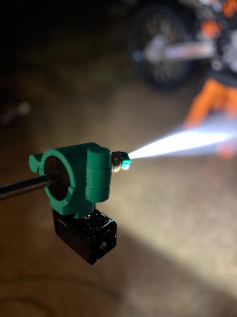 May include: A close-up of a green and black pressure washer nozzle emitting a bright, white stream of water. The nozzle is mounted on a metal rod. The background is dark and out of focus, with a motorcycle visible.