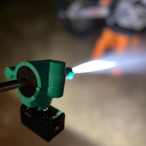 May include: A close-up of a teal-colored torch with a black base, emitting a bright white flame. The torch appears to be made of plastic or a similar material. The background is blurred, suggesting a nighttime setting.