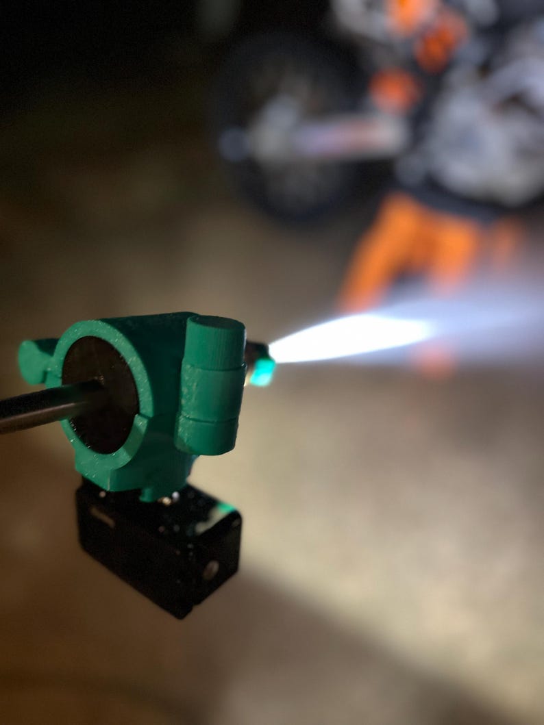 May include: A close-up of a teal-colored torch with a black base, emitting a bright white flame. The torch appears to be made of plastic or a similar material. The background is blurred, suggesting a nighttime setting.