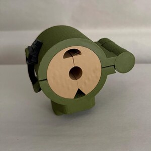 May include: A green, cylindrical object with a tan-colored, circular opening. The object has a rounded top and a smaller, cylindrical extension on the side. A black strap-like detail is visible on the side. The object appears to be made of wood.