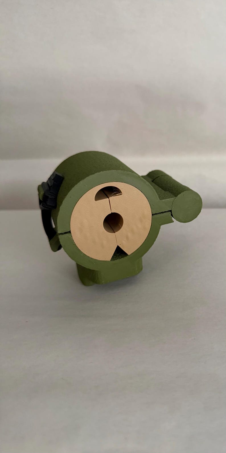 May include: A green, cylindrical object with a tan-colored, circular opening. The object has a rounded top and a smaller, cylindrical extension on the side. A black strap-like detail is visible on the side. The object appears to be made of wood.