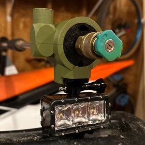 May include: An olive green, 3D-printed device with a brass nozzle and a black light bar. The light bar has three bright LED lights. The device is mounted on a black surface, possibly for outdoor use.
