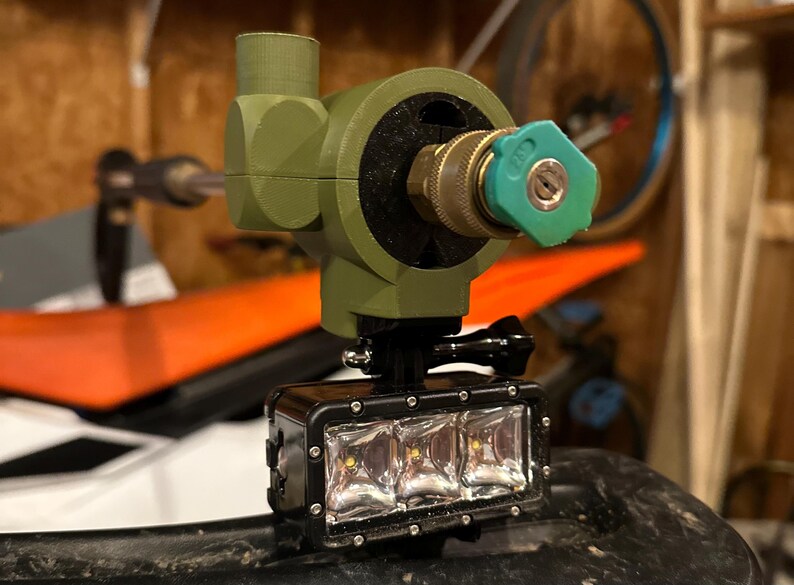 May include: An olive green, 3D-printed device with a brass nozzle and a black light bar. The light bar has three bright LED lights. The device is mounted on a black surface, possibly for outdoor use.