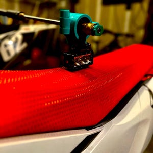 May include: A close-up shot of a teal and black tattoo machine, with a gold-colored nozzle, resting on a red, textured motorcycle seat. The seat is white and black with a number 62 visible in the background.