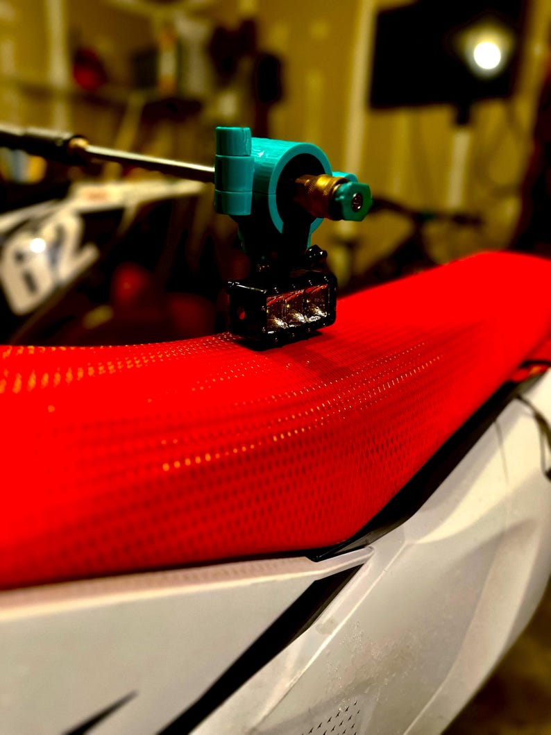 May include: A close-up shot of a teal and black tattoo machine, with a gold-colored nozzle, resting on a red, textured motorcycle seat. The seat is white and black with a number 62 visible in the background.
