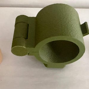 May include: Three 3D-printed objects: a tan cylindrical piece with a patterned top, an olive green cup-shaped item with a handle, and a small black piece. The objects are arranged on a white surface.