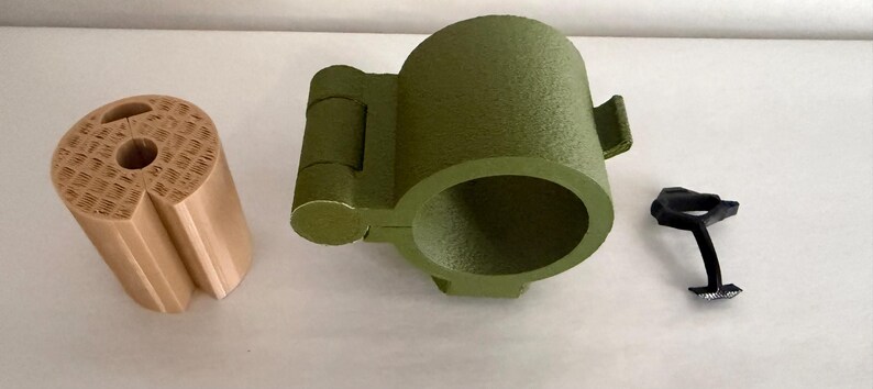 May include: Three 3D-printed objects: a tan cylindrical piece with a patterned top, an olive green cup-shaped item with a handle, and a small black piece. The objects are arranged on a white surface.