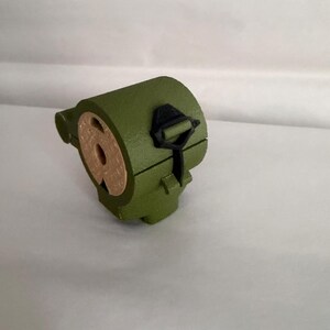 May include: An olive green, cylindrical object with a circular opening and a black clasp. The object has a tan-colored interior and is set against a white background. The design is simple and geometric.