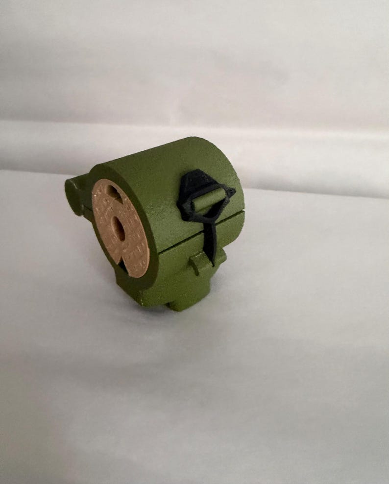May include: An olive green, cylindrical object with a circular opening and a black clasp. The object has a tan-colored interior and is set against a white background. The design is simple and geometric.
