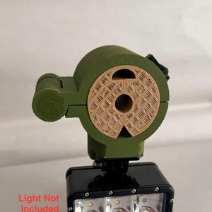 May include: A black and green headlamp mount with a light fixture. The mount is olive green with a tan-colored circular piece. The light fixture has three bright lights. Text on the image reads "Light Not Included."