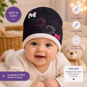 May include: A dark navy blue baby beanie with a white trim, featuring a white letter M, a pink letter N, and two spiral dot patterns in pink and white. The hat includes text labels indicating it is soft, cosy, lightweight, and fits 0-6 months.