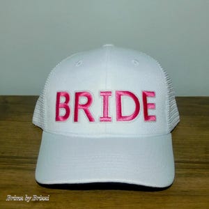 Puede incluir: Gorra de camionero blanca con la palabra "BRIDE" bordada en letras de bloque rosa brillante. La gorra tiene una parte trasera de malla y una visera curva. El texto "Brinz by Brizzi" está impreso en la esquina inferior izquierda.