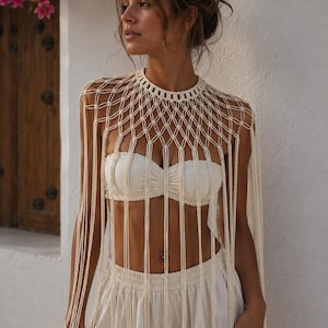 Op de afbeelding: Een gebroken wit macramé capelet met een hoge halslijn en een ingewikkeld geknoopt ruitpatroon. Het kledingstuk is gemaakt van lange, verticale koordstrengen die over de schouders en borst vallen, wat een franje-effect creëert.
