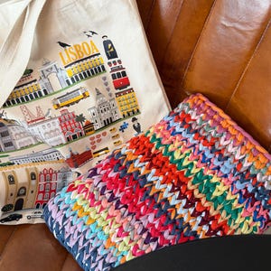 Könnte beinhalten: Eine cremefarbene Canvas-Tragetasche mit einer bunten Illustration von Lissabon-Wahrzeichen und dem Text LISBOA, kombiniert mit einem rechteckigen Kissen aus dickem, mehrfarbig geflochtenem Strickstoff in Rot-, Blau- und Gelbtönen.
