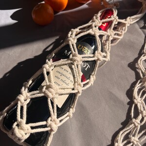 Puede incluir: Una botella de vino tinto en un portabotellas de macram&eacute; color crema. La botella de vino tiene una etiqueta con texto. Naranjas en el fondo. El portabotellas es de cuerda anudada.