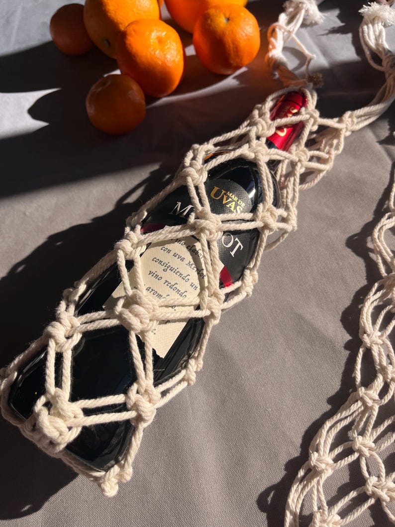 Puede incluir: Una botella de vino tinto en un portabotellas de macram&eacute; color crema. La botella de vino tiene una etiqueta con texto. Naranjas en el fondo. El portabotellas es de cuerda anudada.