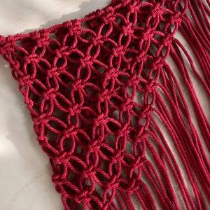 Può includere: Un tessuto in macramè rosso intenso caratterizzato da un motivo a reticolo annodato ripetitivo che forma una serie di forme circolari interconnesse. Il pezzo è rifinito con lunghi fili di frange verticali sciolti che si estendono dai bordi inferiori e laterali.