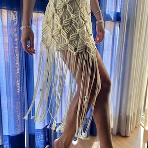 Può includere: Una gonna in macramè color crema caratterizzata da un intricato motivo a rombi annodati nella parte superiore, che sfuma in lunghe frange fluide che scendono fino alla parte inferiore della coscia, rifinite con piccole perline di legno.