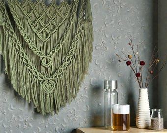 Könnte beinhalten: Olivgrüner Makramee-Wandbehang mit aufwendigen Webmustern und Fransen. Der Textilbehang ist an einem Holzdübel aufgehängt. Die Wanddekoration wird vor einer strukturierten grauen Wand gezeigt. Dekorative Gegenstände auf einem Holztisch.