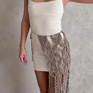 May include: A white sleeveless mini dress paired with a beige macrame waist accessory. The accessory features a knotted diamond pattern with long, dangling fringe that hangs down the side, secured by a thin cord tied around the waist.