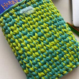 Può includere: Una custodia per libri fatta a mano all'uncinetto, nei toni del verde lime e del verde acqua. La custodia è realizzata con filato spesso e presenta un motivo a punto strutturato. Un libro blu con il titolo "Harry Potter" è parzialmente visibile all'interno. È attaccata una piccola etichetta con il nome "Pryena".