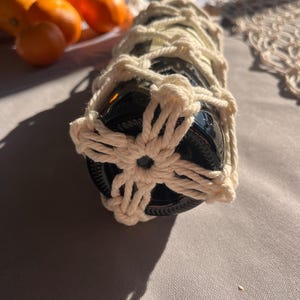 Puede incluir: Una botella de vidrio oscuro envuelta en una red de macram&eacute; color crema. El macram&eacute; presenta un dise&ntilde;o floral en la base. Se ven naranjas en el fondo, lo que sugiere un tema r&uacute;stico o natural.