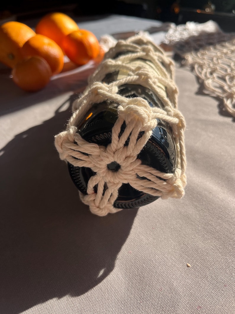 Puede incluir: Una botella de vidrio oscuro envuelta en una red de macram&eacute; color crema. El macram&eacute; presenta un dise&ntilde;o floral en la base. Se ven naranjas en el fondo, lo que sugiere un tema r&uacute;stico o natural.