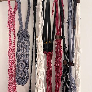 Puede incluir: Una colecci&oacute;n de colgadores de plantas de macram&eacute; hechos a mano en varios colores, incluyendo rojo, gris, negro y blanco, colgados de una barra de madera. Algunos colgadores presentan detalles de cuero y patrones de nudos intrincados, mostrando un estilo bohemio.