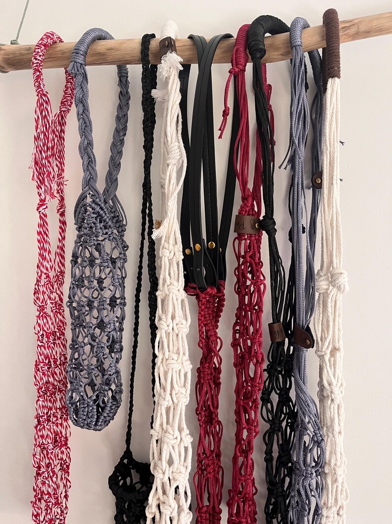 Puede incluir: Una colecci&oacute;n de colgadores de plantas de macram&eacute; hechos a mano en varios colores, incluyendo rojo, gris, negro y blanco, colgados de una barra de madera. Algunos colgadores presentan detalles de cuero y patrones de nudos intrincados, mostrando un estilo bohemio.