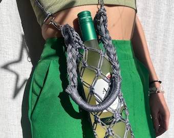 Puede incluir: Un portabotellas de vino de macramé gris con un asa trenzada y una correa para el hombro. El soporte contiene una botella de vino verde. La persona lleva un top y un pantalón verdes.