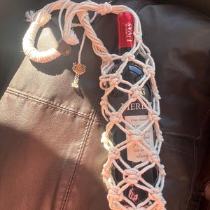 Puede incluir: Un portabotellas de vino de macram&eacute; color crema con un acento de cuero marr&oacute;n y un dije de cuentas. Sostiene una botella de vino tinto oscuro con una etiqueta roja. La botella es parcialmente visible a trav&eacute;s del dise&ntilde;o tejido.