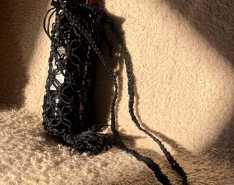 Puede incluir: Un portabotellas de vino de macramé negro con una correa larga y borla. El soporte encierra una botella de vino con una etiqueta roja. El fondo es una superficie texturizada de color marrón claro.