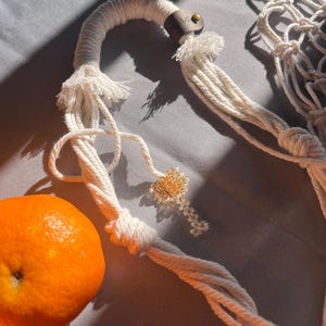 Puede incluir: Primer plano de un colgador de plantas de macram&eacute; blanco con un detalle de cuero marr&oacute;n y un peque&ntilde;o llavero de cuentas. Se ve una fruta naranja. El macram&eacute; est&aacute; sobre una superficie gris.
