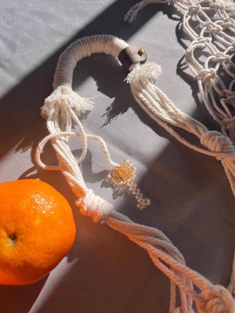 Puede incluir: Primer plano de un colgador de plantas de macram&eacute; blanco con un detalle de cuero marr&oacute;n y un peque&ntilde;o llavero de cuentas. Se ve una fruta naranja. El macram&eacute; est&aacute; sobre una superficie gris.