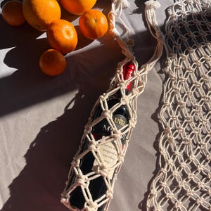 Puede incluir: Un portabotellas de vino de macram&eacute; de color crema, que contiene una botella de vino oscuro con una etiqueta. Naranjas y mandarinas est&aacute;n dispersas cerca. El soporte tiene una correa larga y una cuenta de madera decorativa.