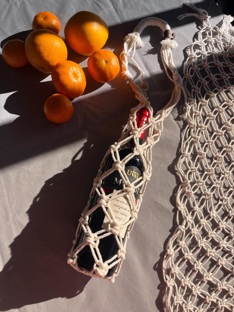 Puede incluir: Un portabotellas de vino de macram&eacute; de color crema, que contiene una botella de vino oscuro con una etiqueta. Naranjas y mandarinas est&aacute;n dispersas cerca. El soporte tiene una correa larga y una cuenta de madera decorativa.