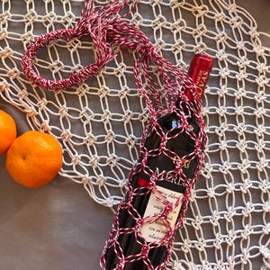 Op de afbeelding: Een rode wijnfles in een macrame wijnfleshouder met rood en wit koord. De houder maakt deel uit van een groter macrame-stuk. Sinaasappels liggen verspreid rond de wijnfles.