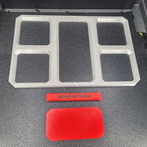 Può includere: Un vassoio organizer rettangolare stampato in 3D di colore bianco con cinque scomparti distinti. Sotto il vassoio si trovano due componenti rettangolari rossi, uno dei quali presenta la scritta nera Springfield Hellcat sulla superficie.