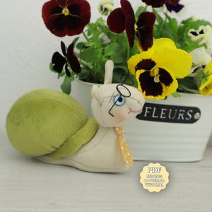 Op de afbeelding: Een pluche slak speelgoed met een groene schaal, een bril en een gele stropdas met stippen. De slak staat voor een witte bloempot met viooltjes. Een klein label leest "PDF Sewing Pattern & Tutorial".