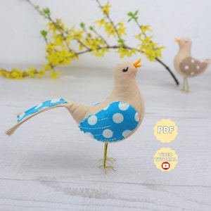May include: A handmade felt bird with a tan body, blue polka-dot wing, and tail. The bird has a yellow beak and delicate gold wire legs. A second bird is in the background. The image includes a PDF and video tutorial icon.