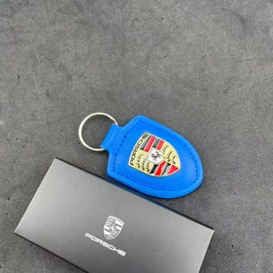 Op de afbeelding: Een felblauwe leren sleutelhanger met een metalen Porsche-embleem in rood, zwart en goud. De sleutelhanger is bevestigd aan een zilverkleurige metalen ring en wordt gepresenteerd naast een zwarte rechthoekige doos met het Porsche-logo in wit.