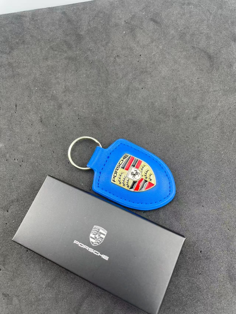 Op de afbeelding: Een felblauwe leren sleutelhanger met een metalen Porsche-embleem in rood, zwart en goud. De sleutelhanger is bevestigd aan een zilverkleurige metalen ring en wordt gepresenteerd naast een zwarte rechthoekige doos met het Porsche-logo in wit.