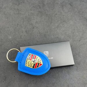 Op de afbeelding: Een felblauwe leren sleutelhanger met een goudkleurig metalen Porsche-embleem met rode en zwarte details. De sleutelhanger is bevestigd aan een ronde goudkleurige metalen ring en ligt naast een kleine zwarte rechthoekige doos.